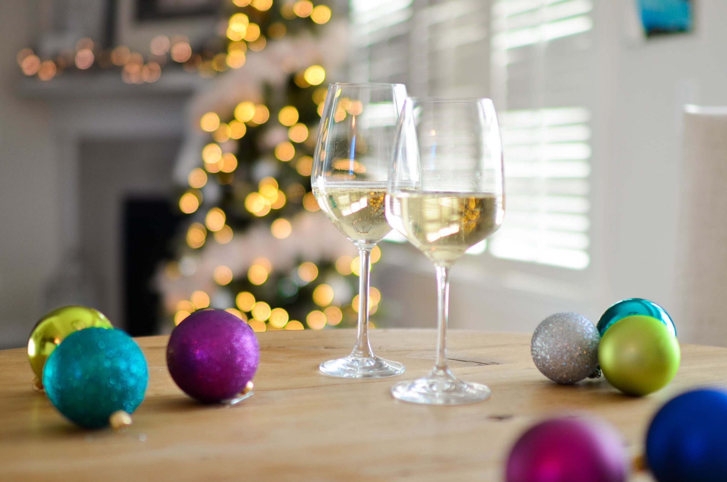 Two glasses of white wine on a wooden table with colourful ornaments and a lit Christmas tree behind