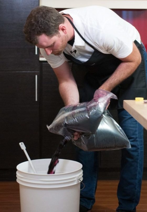 A winemaker in an apron pouring red grape juice from a foil bladder into a white fermenting pail