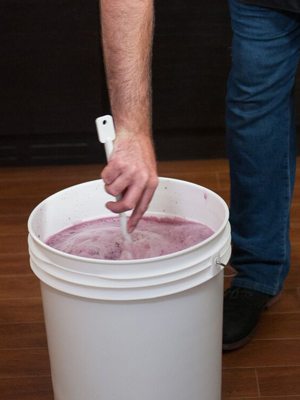A winemaker stirring frothy pink must in a white primary fermenting pail with a long plastic spoon