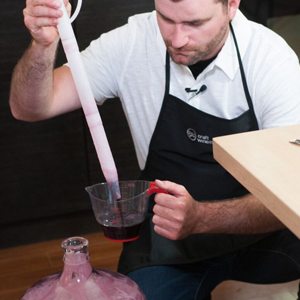 Winemaker holding a long wine thief over a measuring cup of red wine, beside a carboy