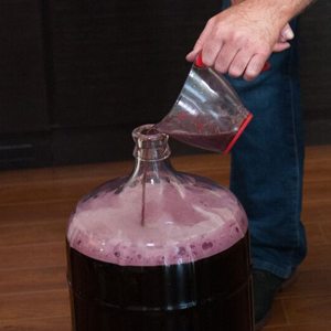 A measuring jug of red wine being poured into the neck of a carboy, raising a pink foam