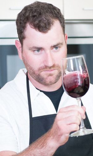 A winemaker in an apron tilting a glass of red wine towards the light to judge its colour and clarity