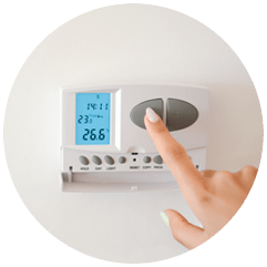 A finger pressing the button on a wall thermostat showing 23 degrees set against 26.6 degrees