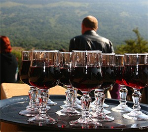 Tray of filled red wine glasses on a table overlooking a forested valley, with a man seated behind